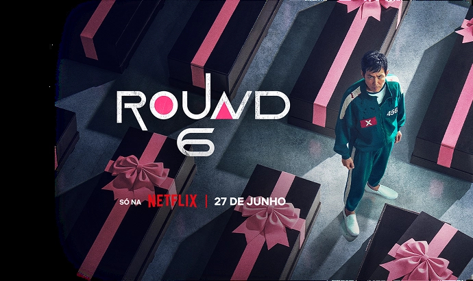 Fundo preto com o texto “Round 6 - Temporada 2” com texto abaixo: “Só na Netflix | Já disponível”. À direita, personagem vestindo um moletom vermelho com capuz e uma máscara preta com um circulo grande no centro.