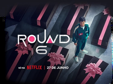 Fundo preto com o texto “Round 6 - Temporada 2” com texto abaixo: “Só na Netflix | Já disponível”. À direita, personagem vestindo um moletom vermelho com capuz e uma máscara preta com um circulo grande no centro.