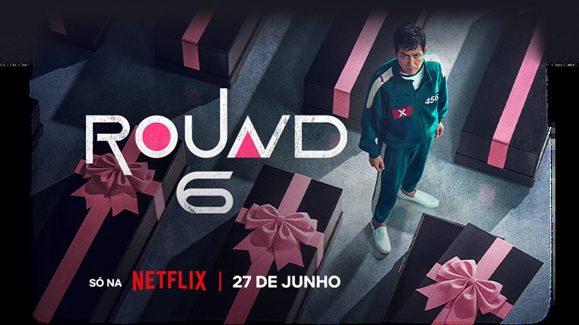 Fundo preto com o texto “Round 6 - Temporada 2” com texto abaixo: “Só na Netflix | Já disponível”. À direita, personagem vestindo um moletom vermelho com capuz e uma máscara preta com um circulo grande no centro.