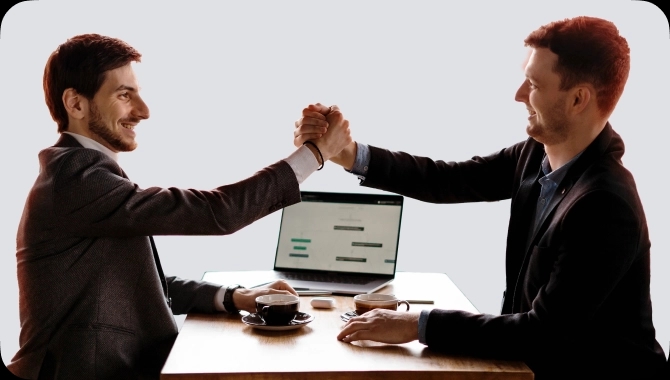 Dois homens em um ambiente de trabalho, vestindo ternos, sentados frente a frente em uma mesa com duas xícaras de café e um laptop ao fundo. Eles estão sorrindo e apertando as mãos. 