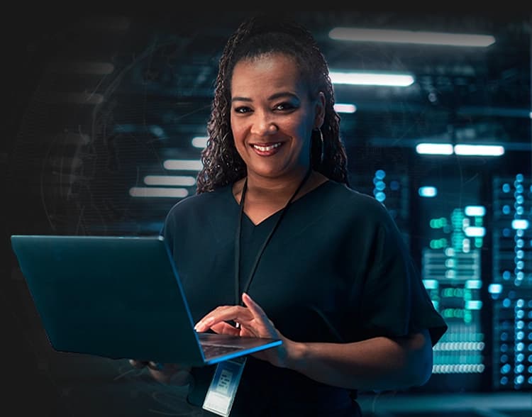 Uma mulher segurando um notebook aberto. Ela veste uma blusa escura e usa um crachá pendurado no pescoço. Ao fundo, é exibido um ambiente de sala de servidores com corredores iluminados
