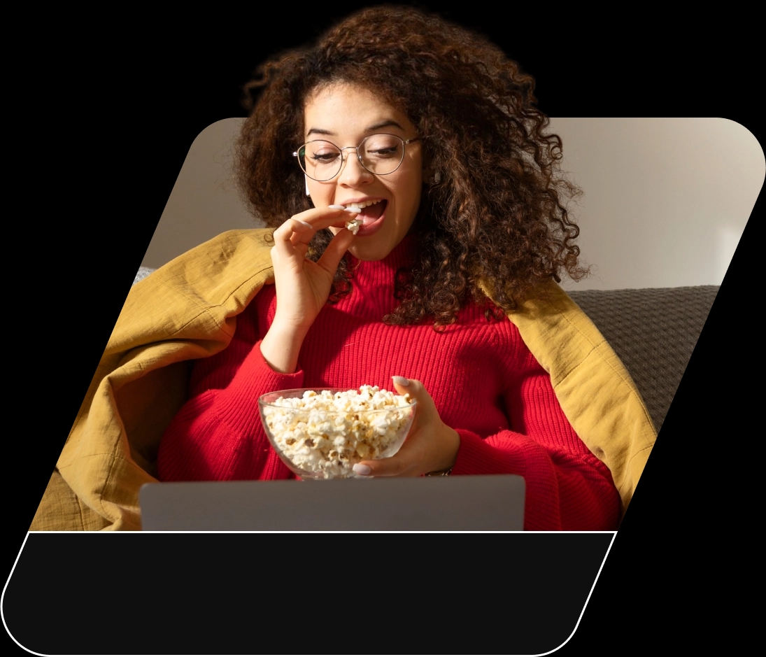 Mulher de pele branca e cabelos cacheados sentada no sofá comendo pipoca assistindo série em seu notebook