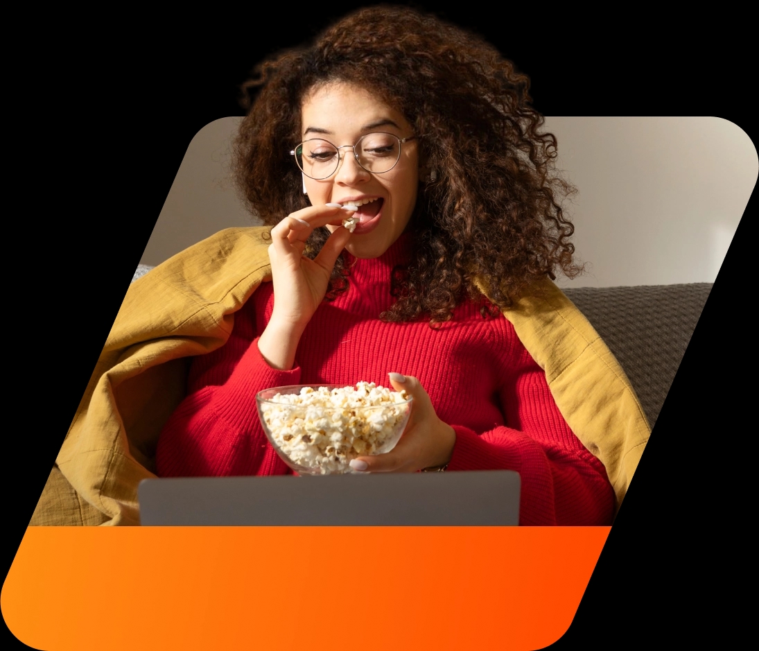 Mulher de pele branca e cabelos cacheados sentada no sofá comendo pipoca assistindo série em seu notebook